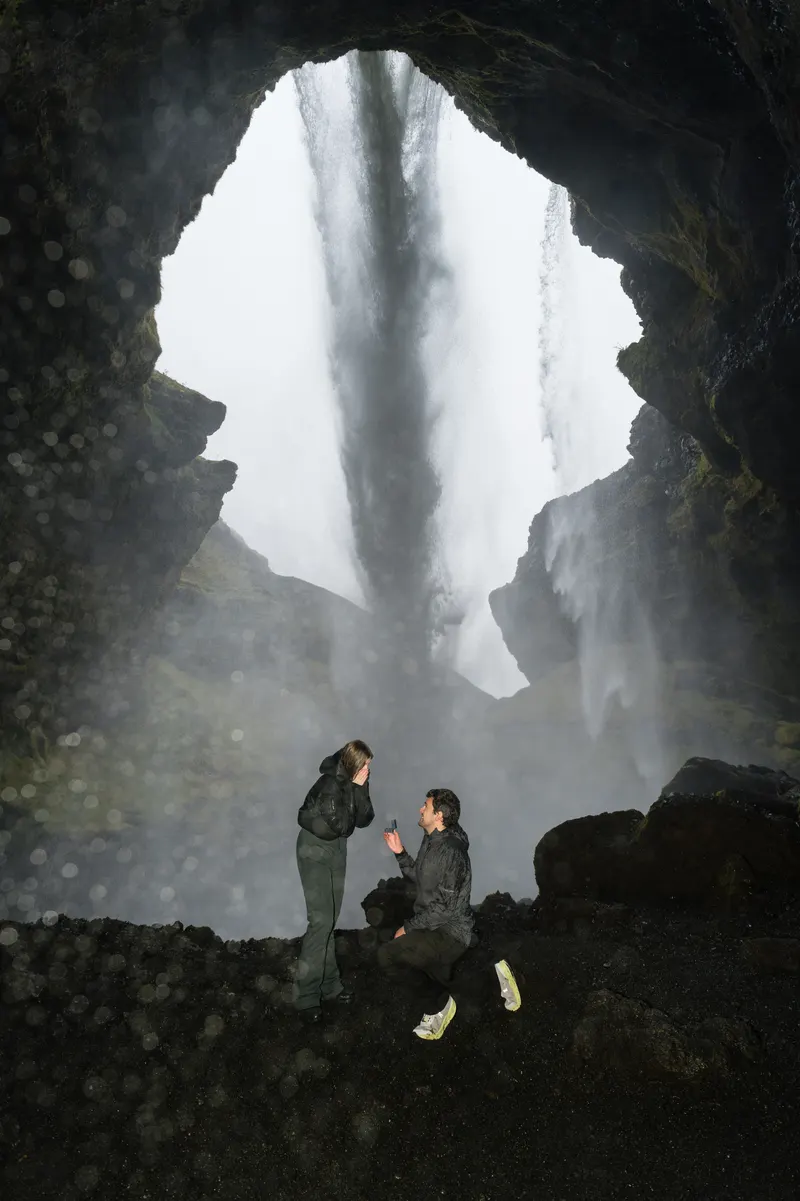 Edu, de rodillas sobre la roca negra, le pide matrimonio a Dani en la cueva que hay detrás de la cascada de Kvernufoss; ella se lleva las manos a la cara.